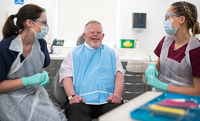 A man sitting in a dentist's chair chats to a dentist and her assistant